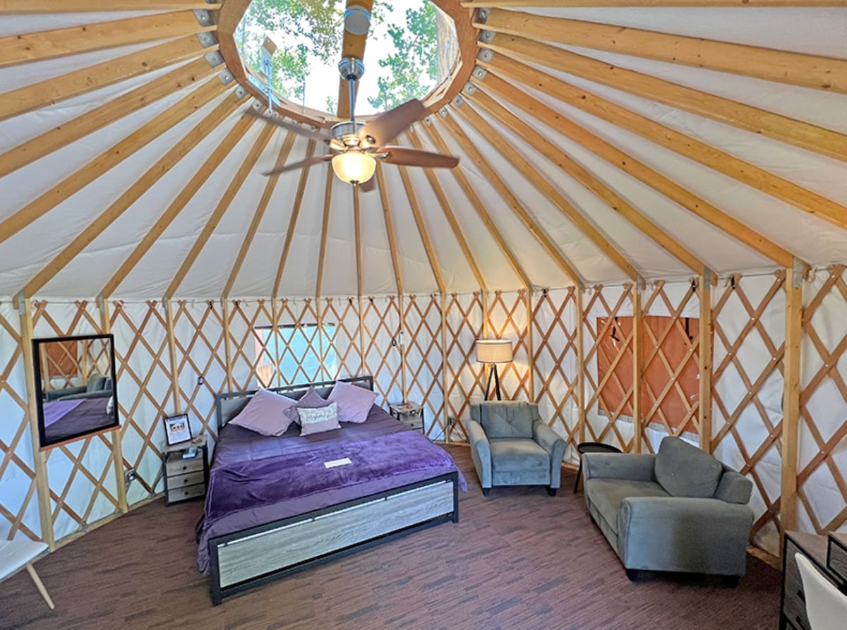 The interior of the yurt features a spacious bed with purple bedding, complemented by two cozy chairs. Natural light filters through a circular skylight at the center of the wooden-beamed ceiling, enhancing the airy feel of the room. A mirror and bedside tables are visible.