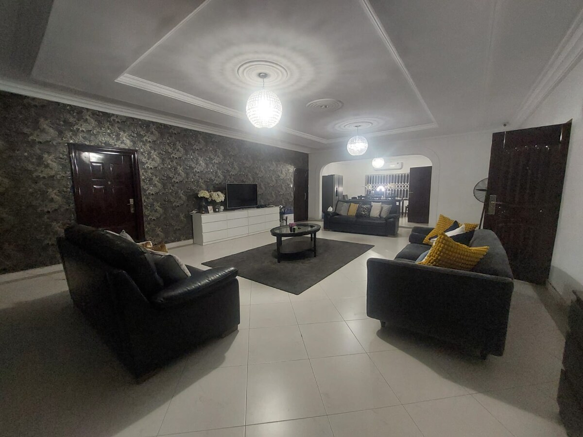 A spacious living room is showcased, featuring two dark sofas and a glass coffee table on a large area rug. Soft lighting is provided by modern chandeliers. A stylish entertainment unit and an arched entryway lead to other areas, enhancing the room's elegance.