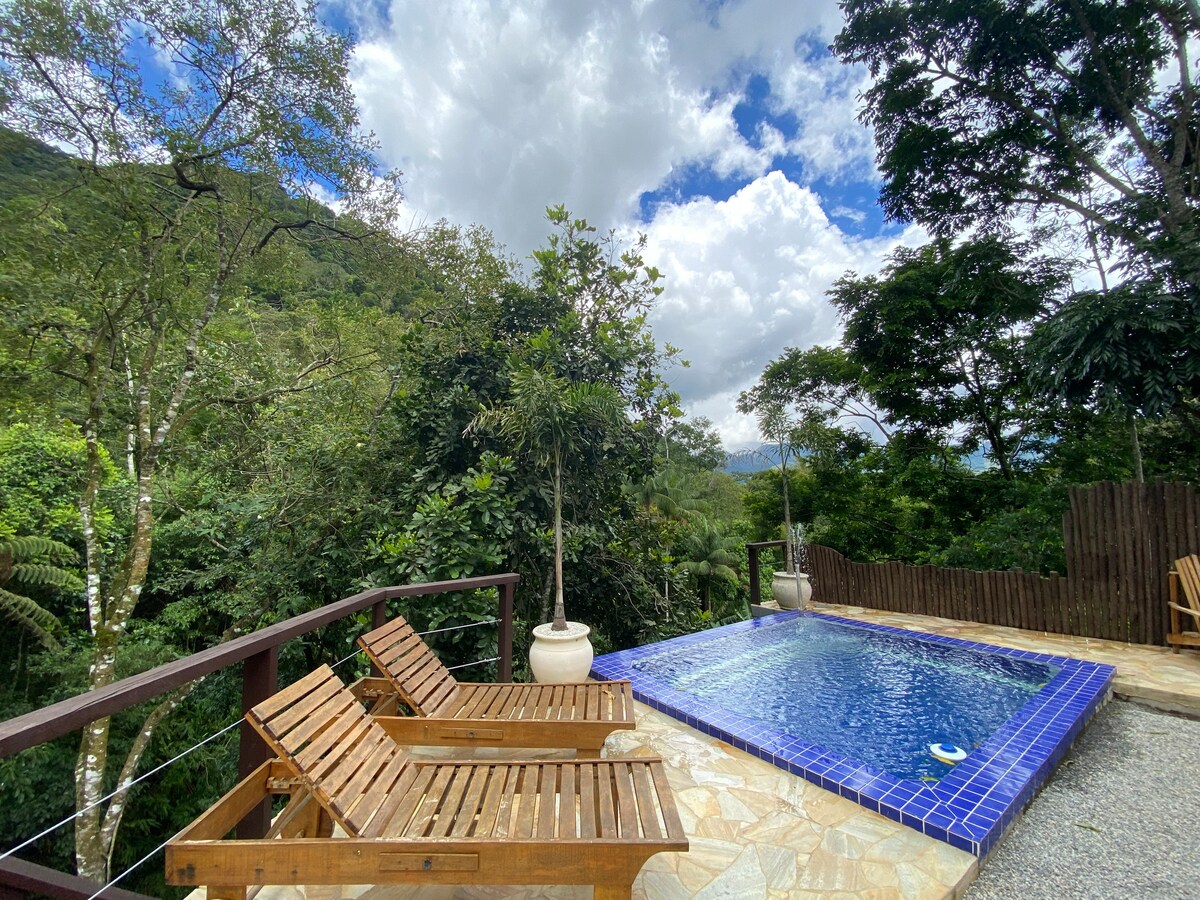A serene private area features a blue-tiled pool surrounded by lush greenery. Two wooden lounge chairs provide seating options. The landscape is accentuated by a variety of trees and distant hills under a partly cloudy sky.
