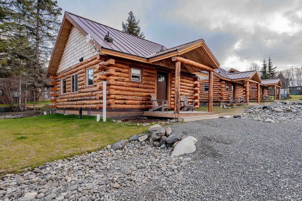A cluster of rustic log cabins sits under a cloudy sky. Each cabin features a covered porch with seating, and the landscape is framed by lush grass and strategically placed stones, contributing to a natural setting along the shore.