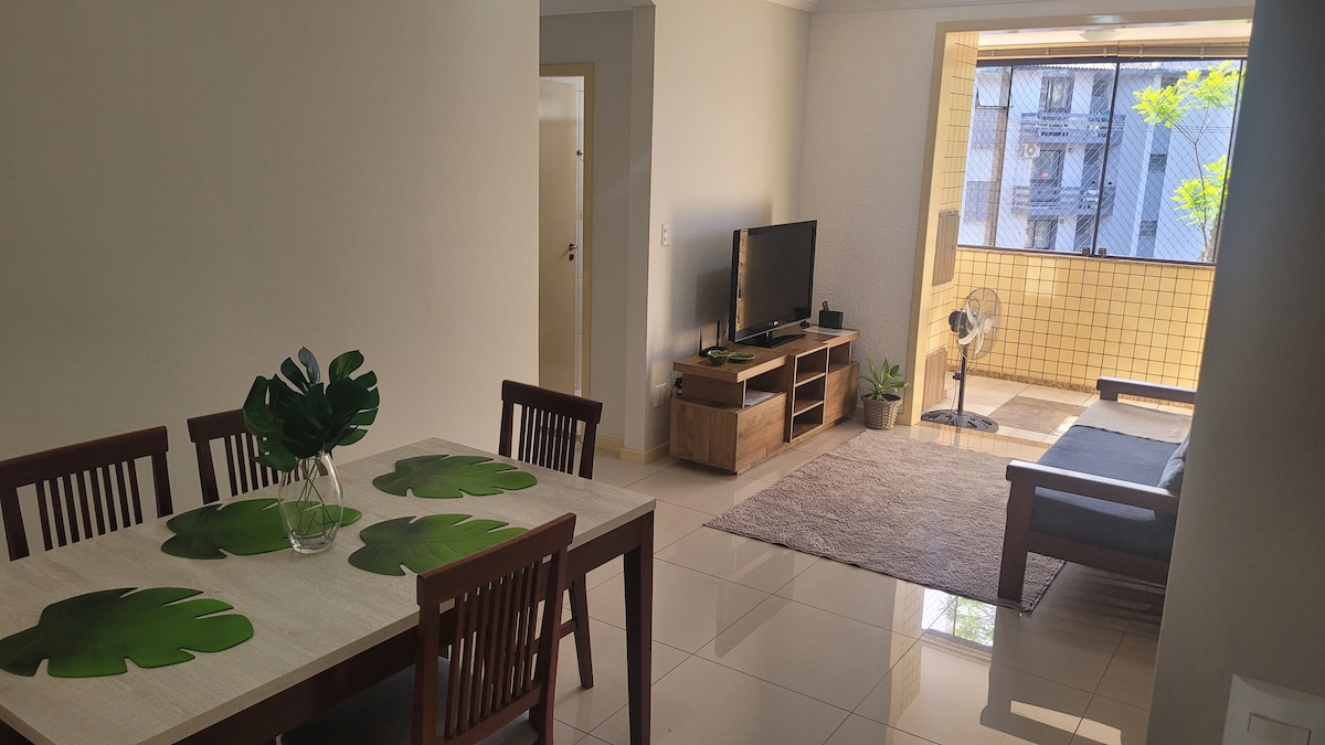 A spacious living area features a dining table with leaf-patterned placemats, complemented by a comfortable sofa and a fan. Natural light filters in through a large window, offering a view of greenery and creating a bright and airy atmosphere.