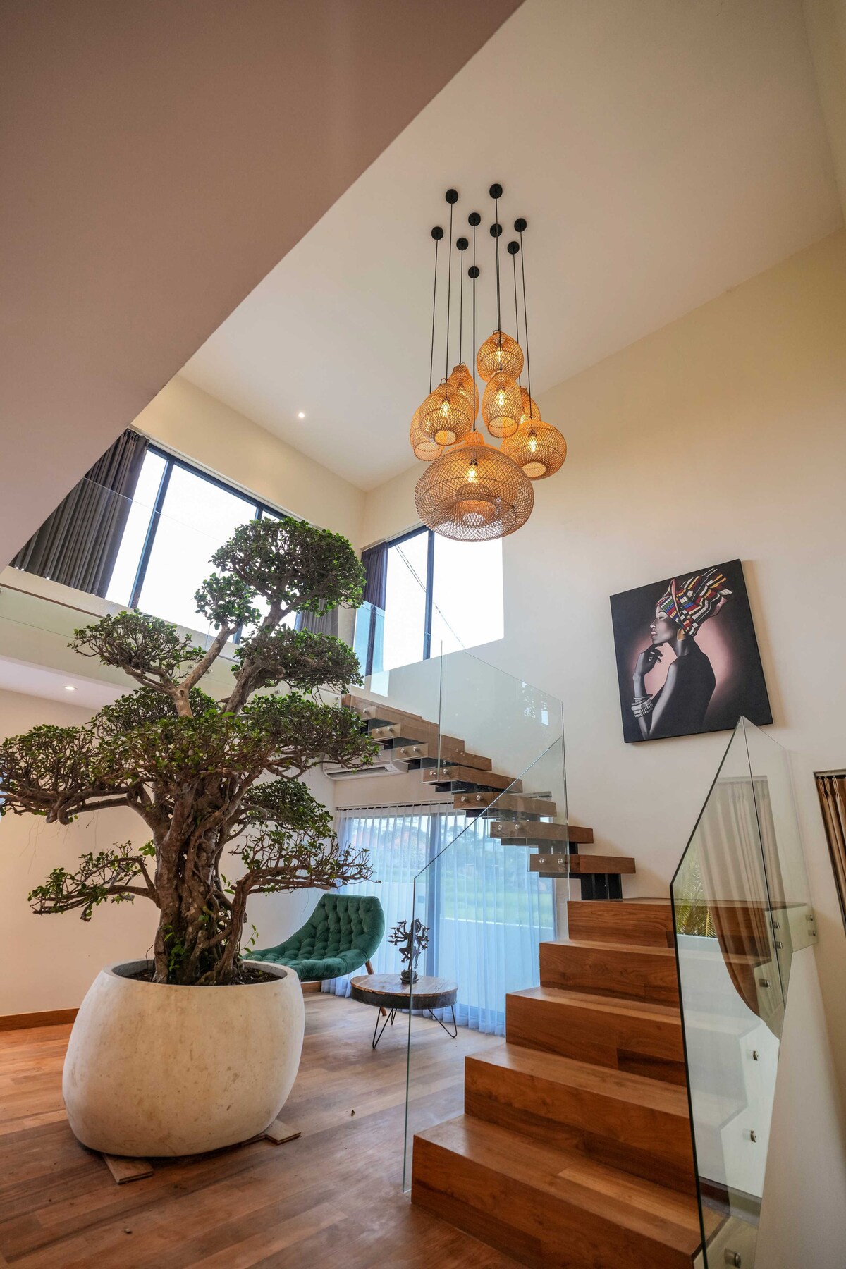 A well-lit staircase features wooden steps and a glass railing, leading to an upper level. A large potted tree adds a natural element, while modern hanging lights create a warm ambiance. Artwork adorns the wall, complementing the contemporary design of the space.
