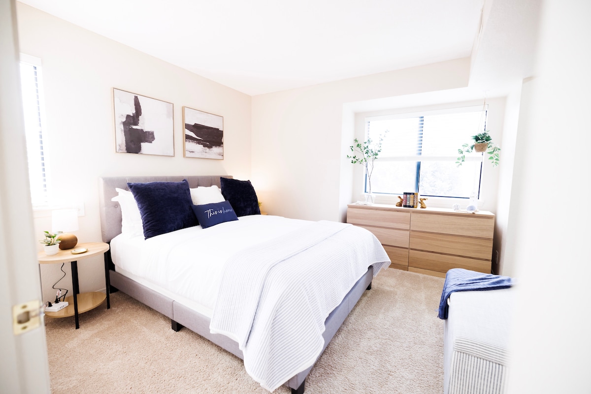 A serene bedroom features a spacious bed adorned with a white blanket and navy accent pillows. Two pieces of artwork hang above the bed, while natural light filters through the two windows. A wooden dresser and bedside table complement the neutral color palette.