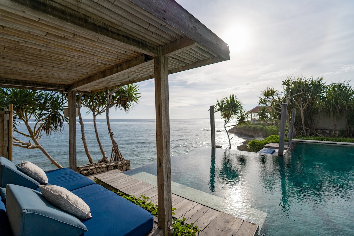A stylish lounge area is presented under a wooden structure, offering comfortable blue cushions. The stunning view includes a shimmering infinity pool which merges with the ocean horizon, framed by lush tropical greenery and a clear sky.
