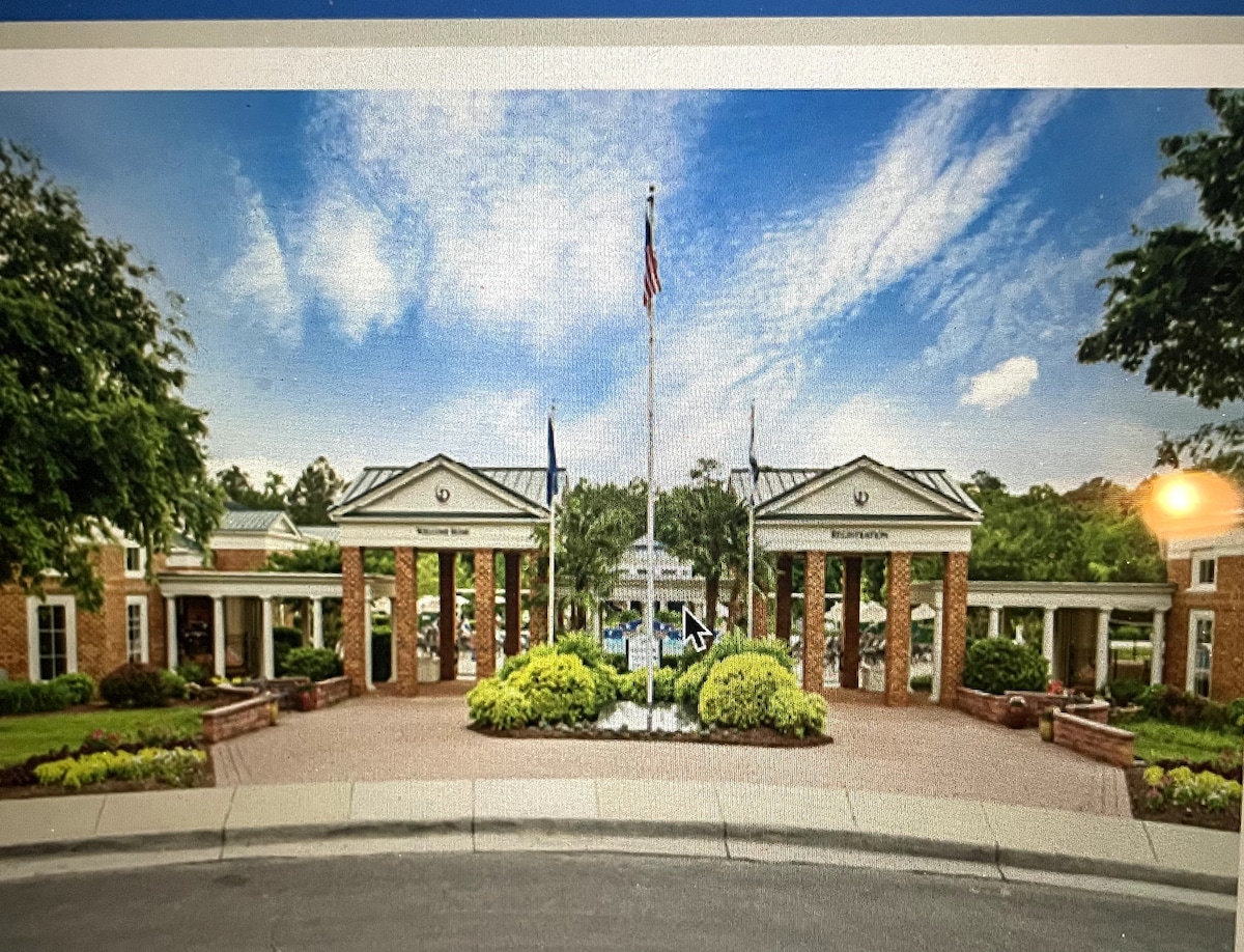An entrance with a welcoming design features brick structures flanking a central path. Lush greenery surrounds the area, including neatly trimmed bushes and flower beds. Flags are displayed, and the blue sky above is dotted with white clouds, adding to the inviting atmosphere of the property.