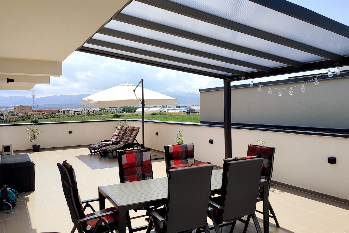 A spacious terrace is shown, featuring a dining table surrounded by six chairs. Two lounge chairs with cushions are positioned nearby, under a large umbrella. Clear views of the surrounding landscape and mountains are visible in the background.