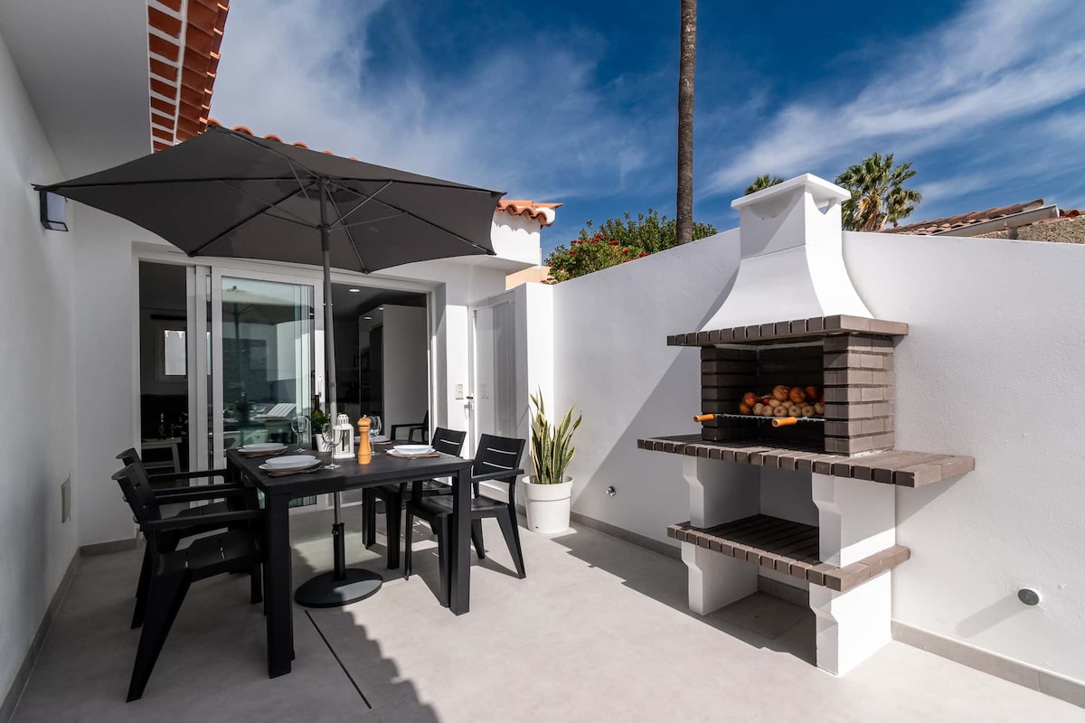 A private patio features a dining table with four chairs and a large umbrella for shade. A built-in barbecue stands nearby, with neatly arranged logs for grilling. The space is enclosed by walls and is illuminated by natural light.