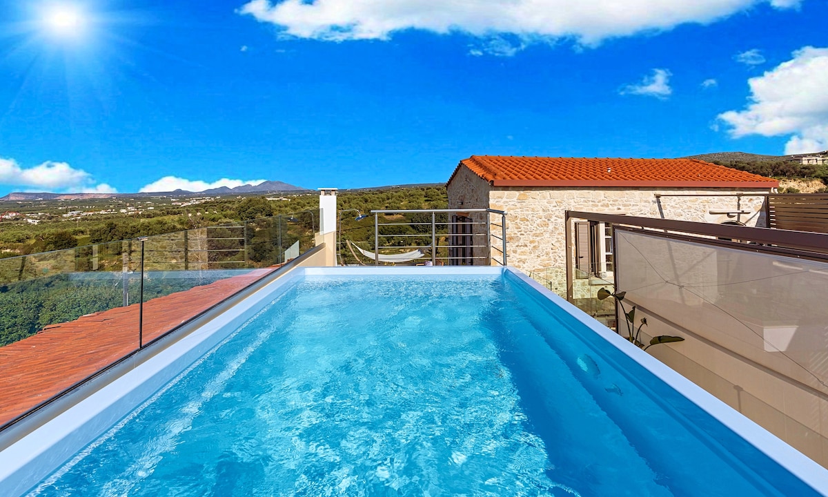 A heated whirlpool is situated on a terrace overlooking a scenic landscape, featuring a combination of stone buildings and greenery. The clear blue sky and bright sunlight enhance the tranquil atmosphere. The surrounding area reflects the natural beauty of Crete.