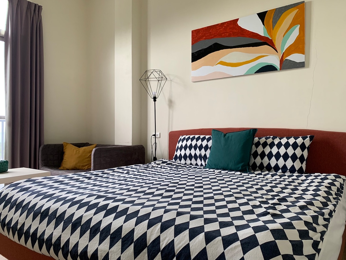 A spacious bed is covered with a patterned checkered blanket in black and white. A decorative pillow in green is placed against a colorful wall art piece above. A modern floor lamp stands beside the bed, with natural light filtering in through a large window.