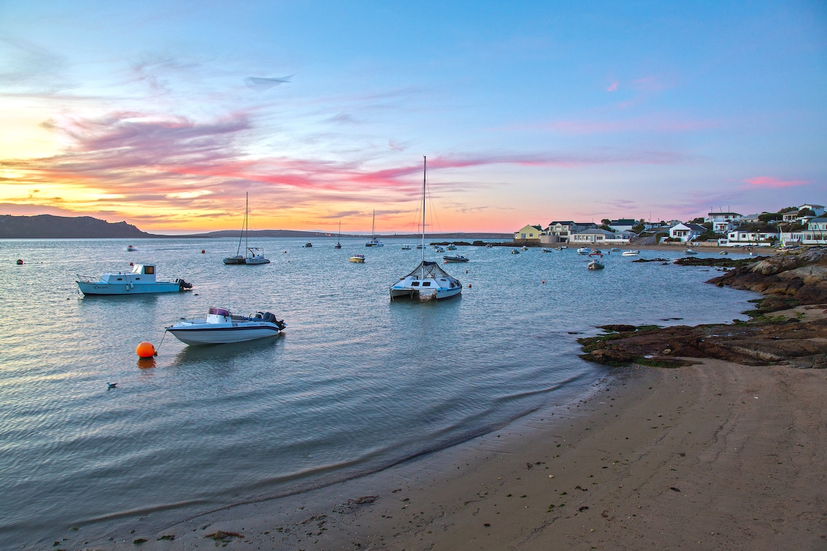 The image captures a serene coastal scene during sunset, with several boats gently floating in the calm water. The shoreline is sandy and features a mix of rocky outcrops, while pastel colors from the sky reflect on the surface of the water.