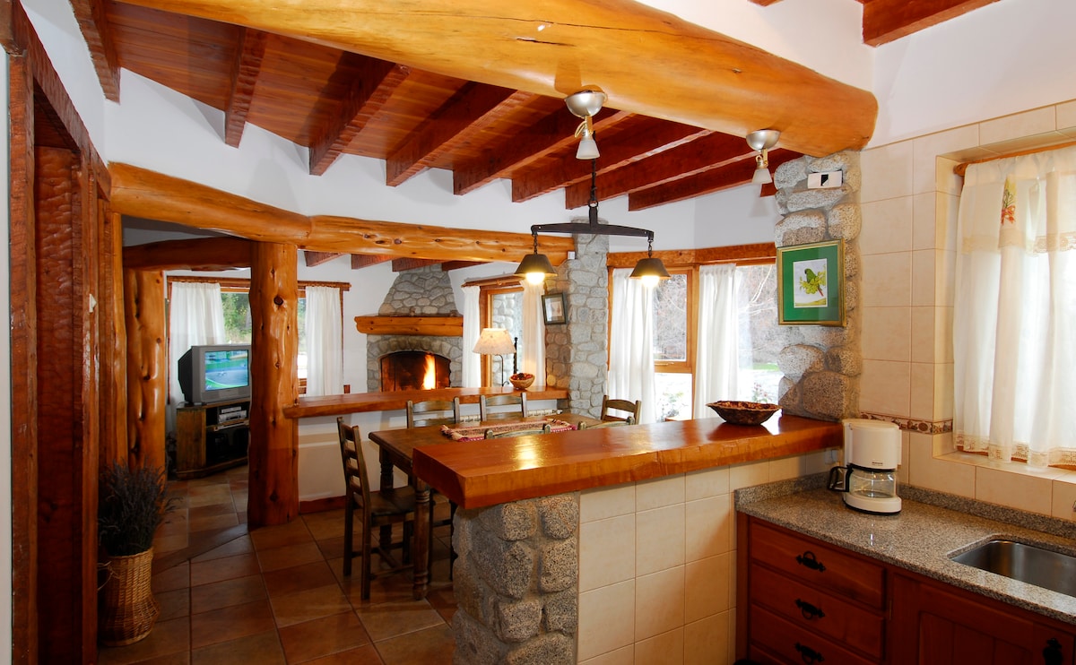 A welcoming kitchen and living area feature rustic wooden beams and stone accents. Natural light filters through large windows, illuminating the cooking space, which includes a coffee maker and generous countertop. A cozy seating arrangement and a fireplace create a warm focal point in the room.