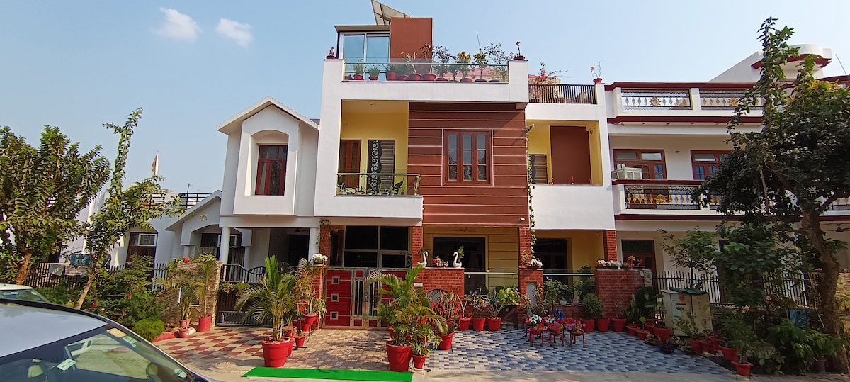 The villa's exterior displays a blend of modern and traditional architecture, featuring multiple floors with a terrace adorned with potted plants. A vibrant garden with red flower pots flanks the entrance, and a spacious driveway is visible for vehicle access.