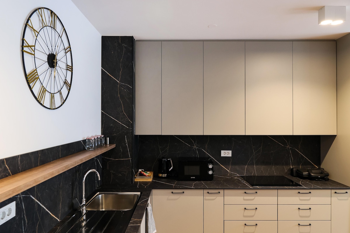 A sleek kitchen is presented with dark marble-patterned walls and modern tan cabinetry. A wooden shelf extends along one wall, displaying glassware. A stainless steel sink is positioned under a decorative clock, with a microwave and stovetop visible on the counter.