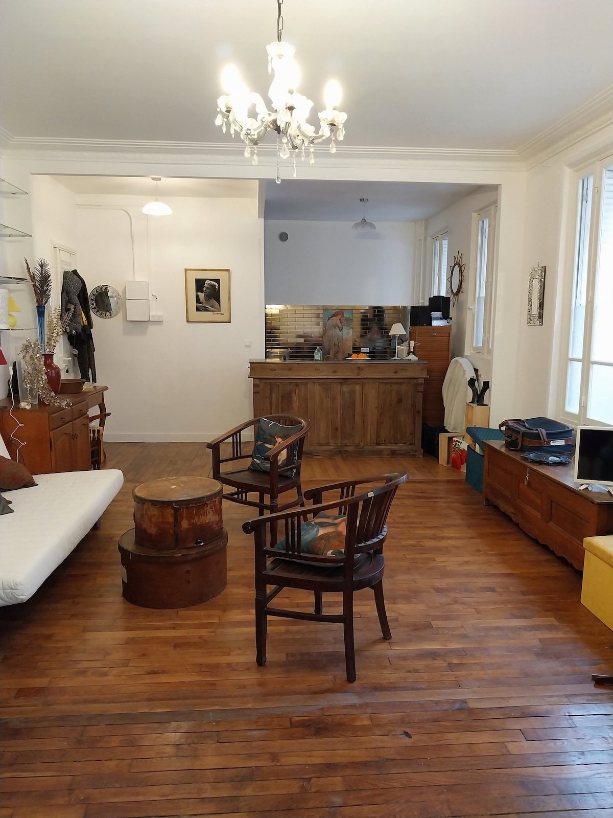 An inviting living area features warm wooden flooring and a blend of vintage furniture. Two wooden chairs are positioned around a circular table, with a convertible sofa to one side. A charming chandelier hangs from the ceiling, and a kitchen area is visible in the background.