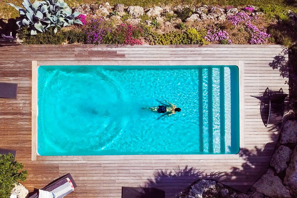 An aerial view captures a clear swimming pool surrounded by wooden decking and lush greenery. Vivid flowers line the edges, adding color to the setting. A swimmer can be seen enjoying the water, emphasizing the pool's inviting nature.