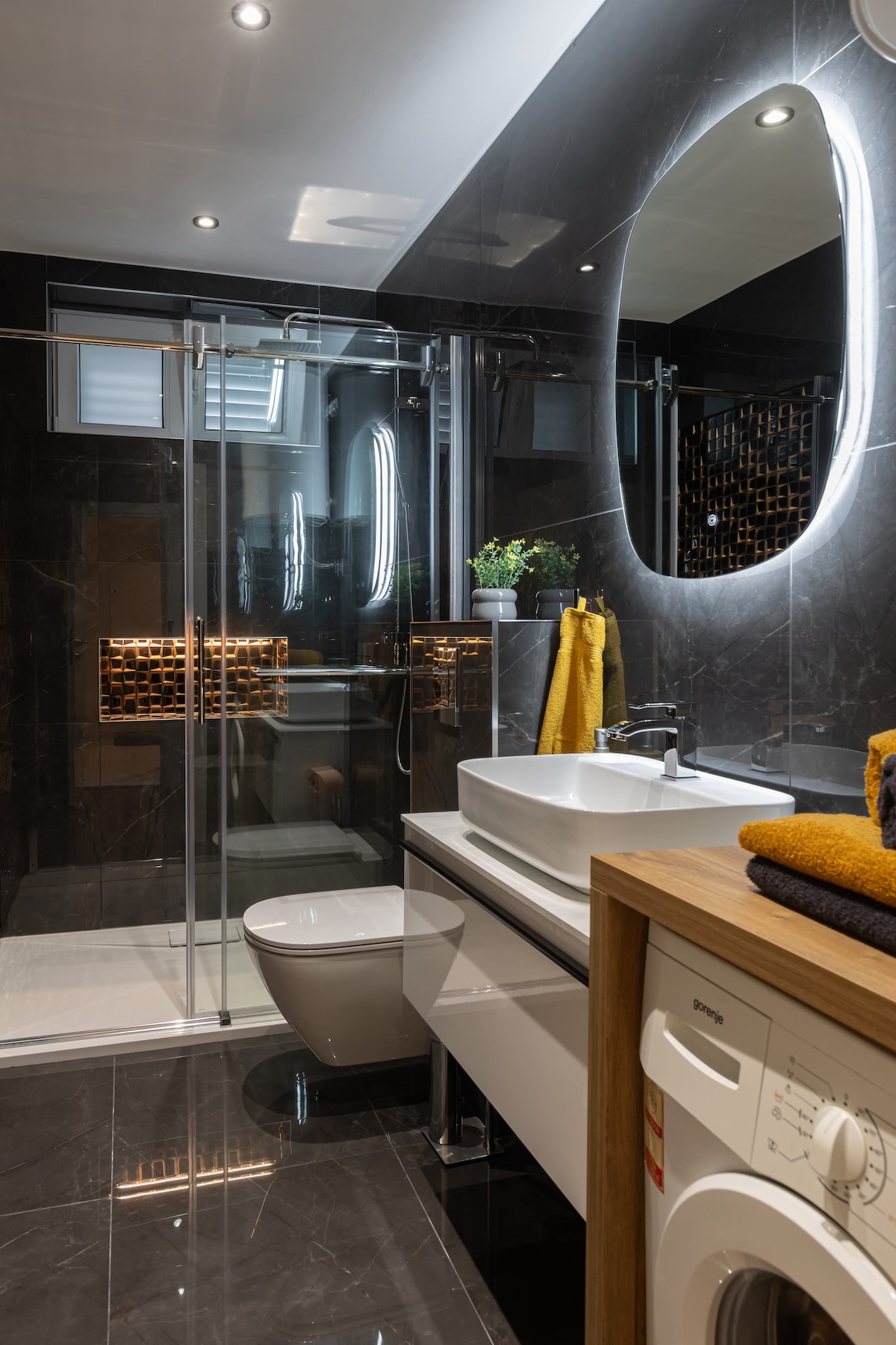 A modern bathroom features sleek black tiles and a spacious glass shower. A large illuminated mirror is mounted above a white sink. A compact washing machine sits under a wooden countertop, complemented by neatly arranged towels and a decorative plant.