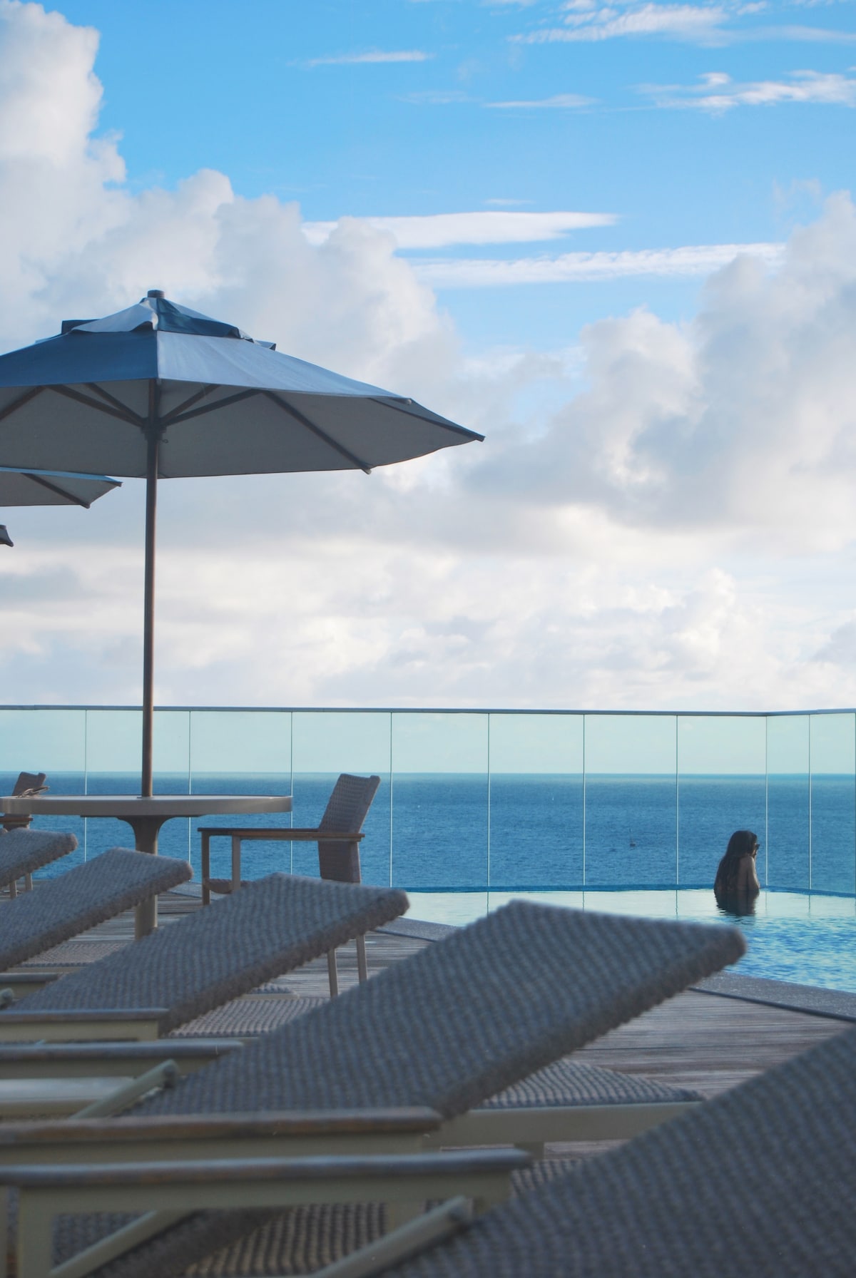 A tranquil infinity pool is featured, overlooking the sea under a bright sky. Lounge chairs are arranged beside the pool, and a shaded table with an umbrella is visible nearby. Soft clouds enhance the serene atmosphere.