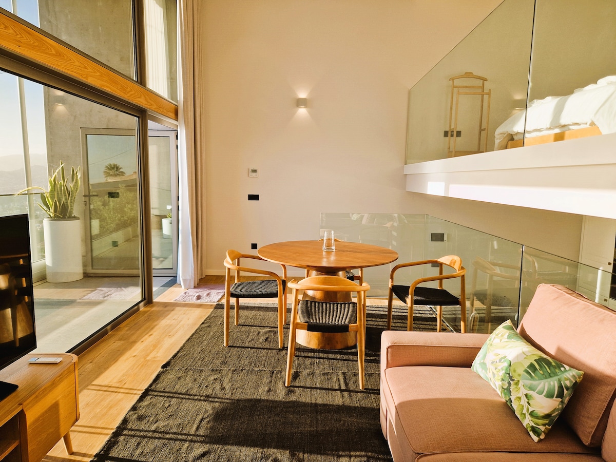 A modern living area is highlighted by a circular wooden dining table surrounded by four chairs. A comfortable armchair with a floral pillow is positioned near a glass wall, allowing natural light to fill the space and offering views of the outdoors.