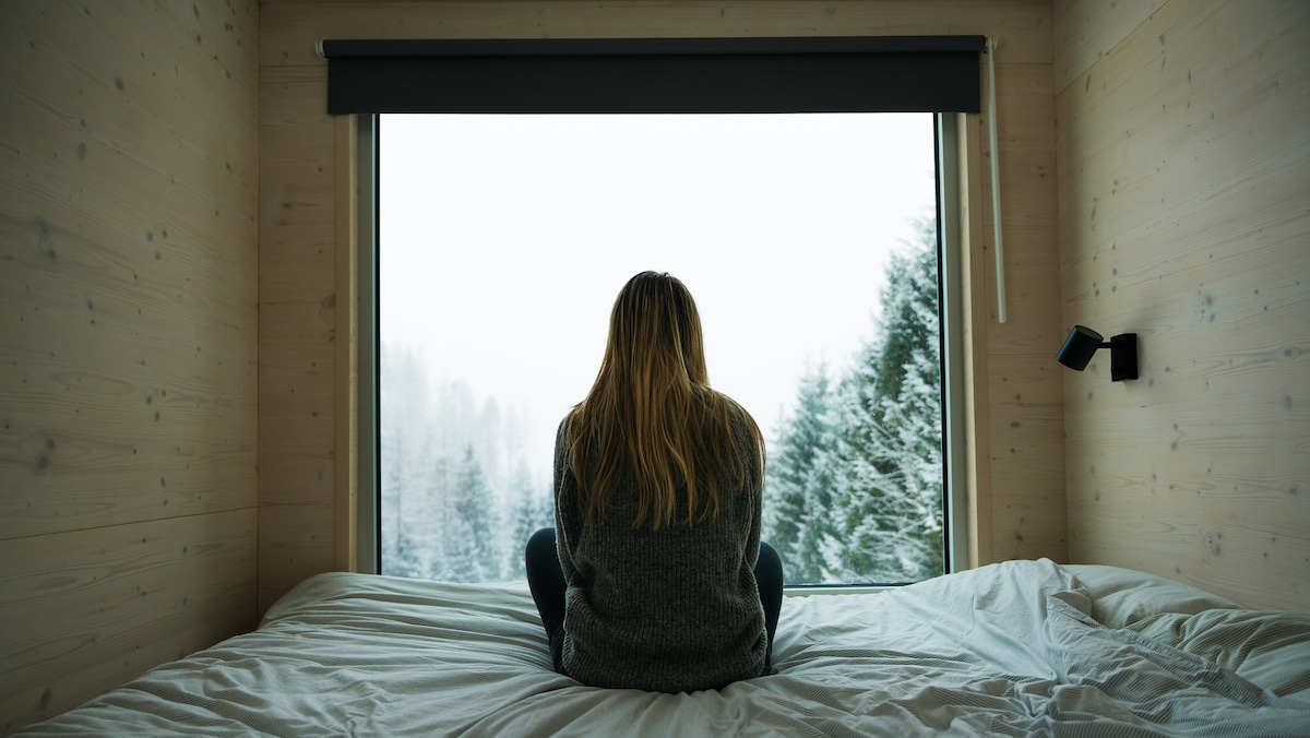 A person sits on a spacious bed, facing a large window that frames a snowy landscape. The soft natural light filters through, highlighting the serene environment of trees blanketed in snow. The wood-paneled walls add warmth to the minimalist space.