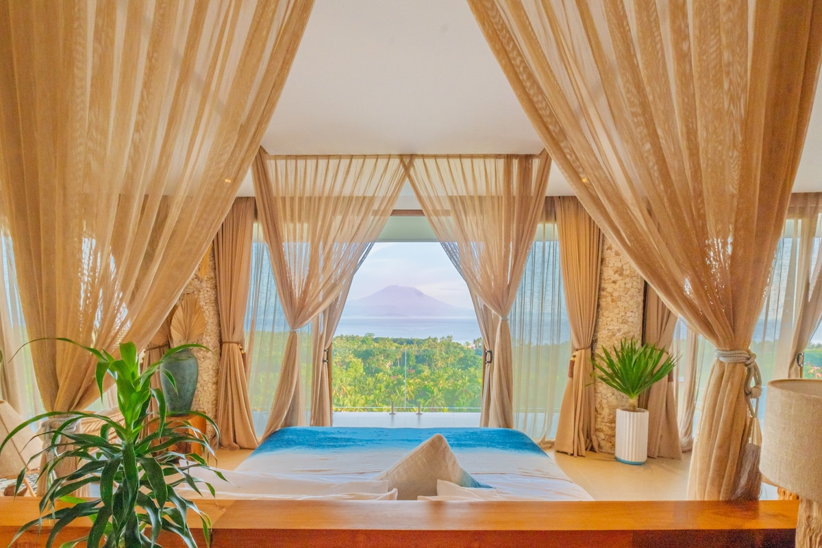 A spacious bedroom is highlighted by flowing sheer curtains framing a large window. A calming view of lush greenery and distant hills is visible beyond the glass. A decorative plant adds a touch of nature to the room, enhancing the overall serene ambiance.