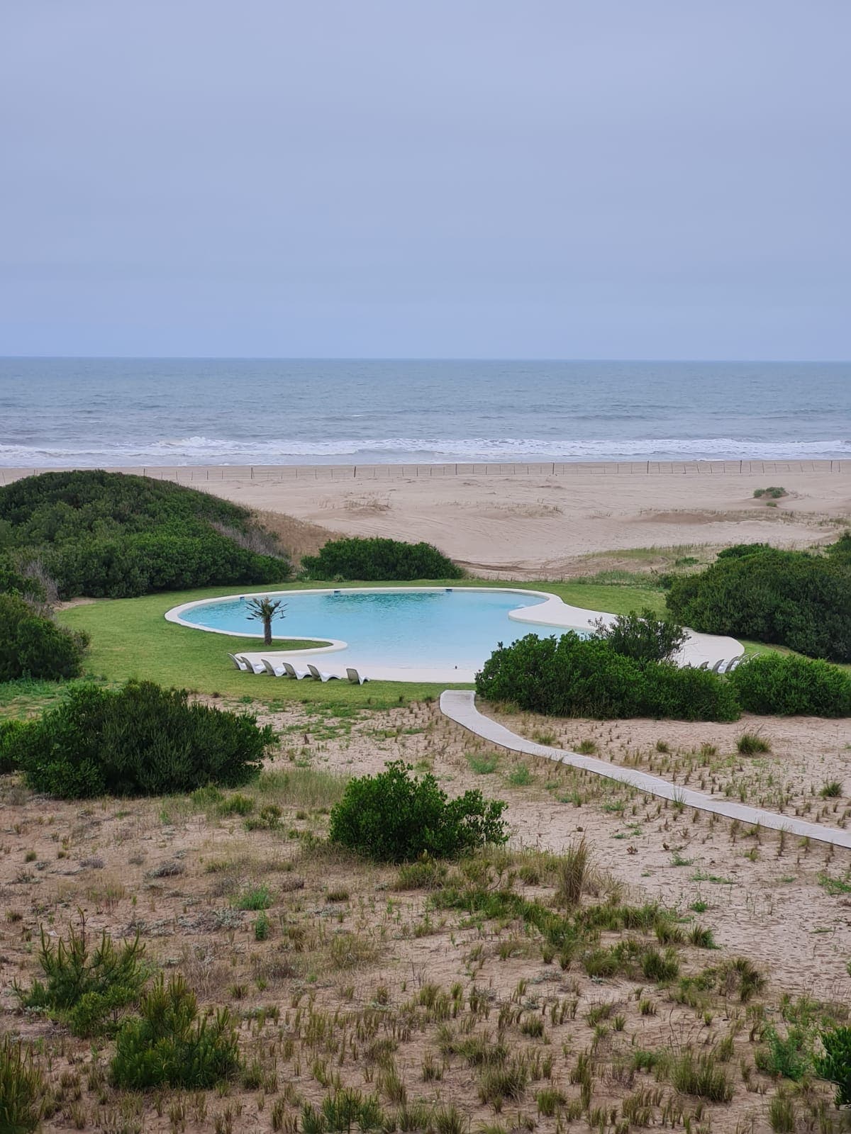 An outdoor pool is framed by lush greenery, offering a serene view of the sandy beach and ocean beyond. A winding path leads from the pool area to the beach, creating a seamless connection between the two. The water reflects the cloudy sky.