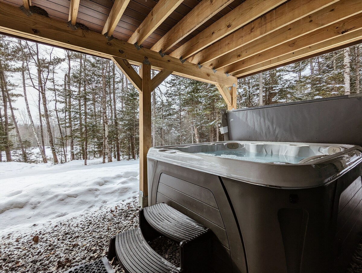 A hot tub is positioned under a wooden shelter, surrounded by trees and snow-covered ground. Steps lead up to the hot tub, providing easy access. The tranquil setting offers views of the forest, enhancing the sense of privacy and relaxation.