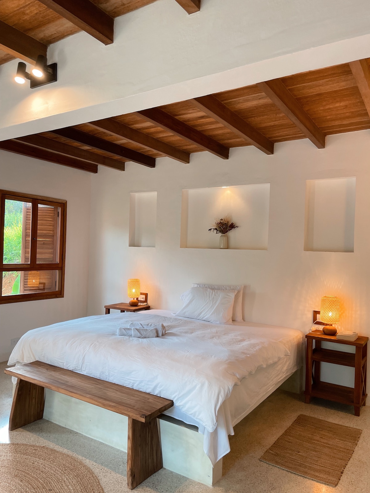 A spacious bedroom features a large bed with crisp white linens, complemented by wooden side tables with subtle lighting. A low wooden bench sits at the foot of the bed, and a window permits natural light while offering a view of the surrounding greenery.