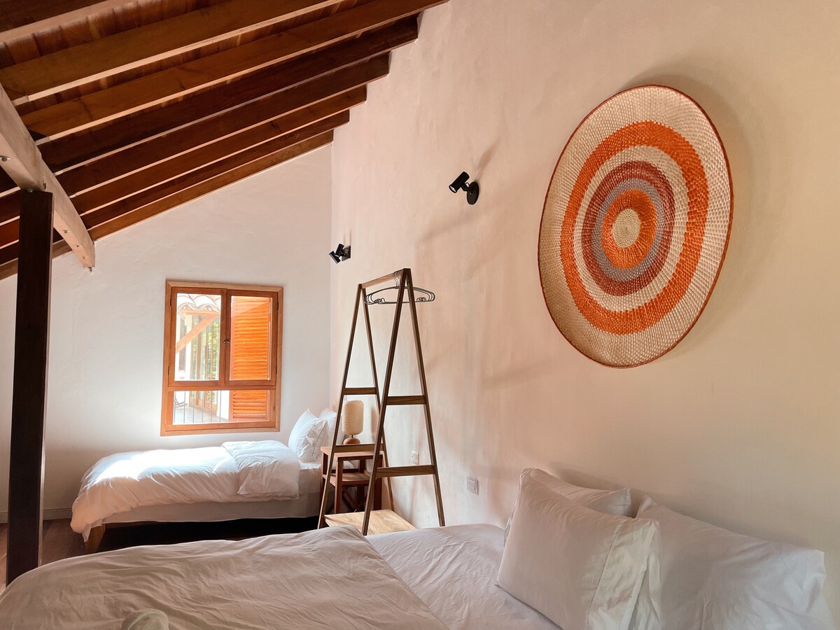 A cozy room features two beds with white linens, complemented by wooden beams on the ceiling. A circular woven wall art adorns one wall, while a ladder-style shelf and a chair provide functional decor. A window offers natural light and a view of the outdoors.