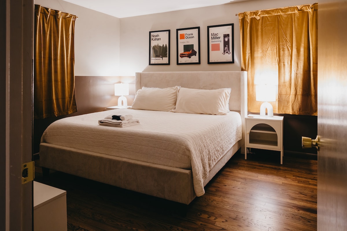 A serene bedroom features a spacious bed with soft white linens and two side tables. Warm golden curtains frame the window, and contemporary artwork adorns the walls. Soft lighting from bedside lamps creates a calming ambiance, inviting relaxation for restful nights.