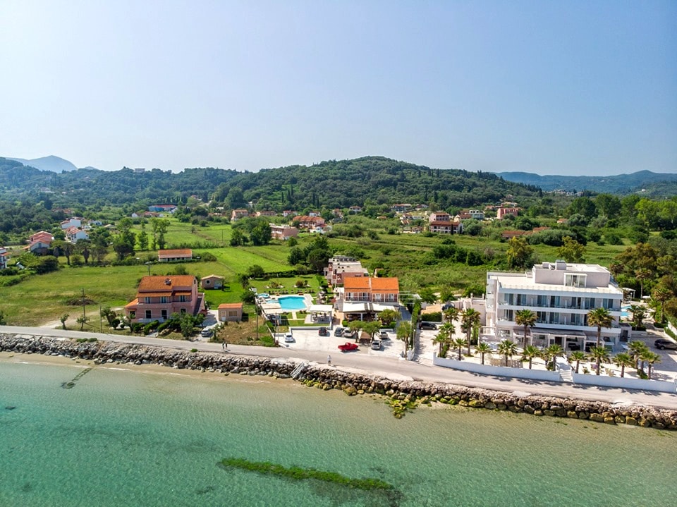 An aerial view captures a coastal area featuring several buildings, including a two-story structure. A large pool is visible, surrounded by green lawns. The serene sea lies in the foreground, with sandy shores blending into tranquil waters, and lush greenery extending into the background.