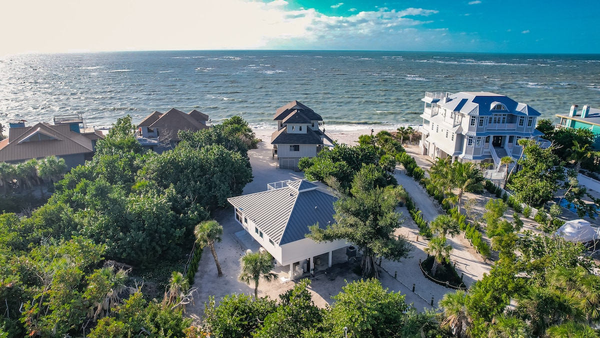 An aerial view of a beach house surrounded by lush greenery. The ocean is visible in the background, reflecting the sunlight. The home's rooftop features a crow's nest, and multiple neighboring houses can be seen along the shoreline.