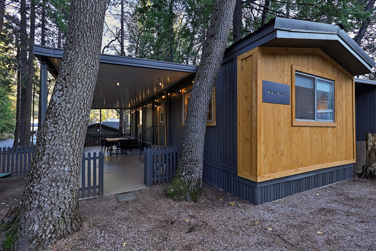 A cabin exterior features a mix of wooden and gray siding, surrounded by tall trees. A patio area is seen, with a fence providing separation. Warm lighting enhances the inviting atmosphere, while a sign displays the cabin's name near the entrance.