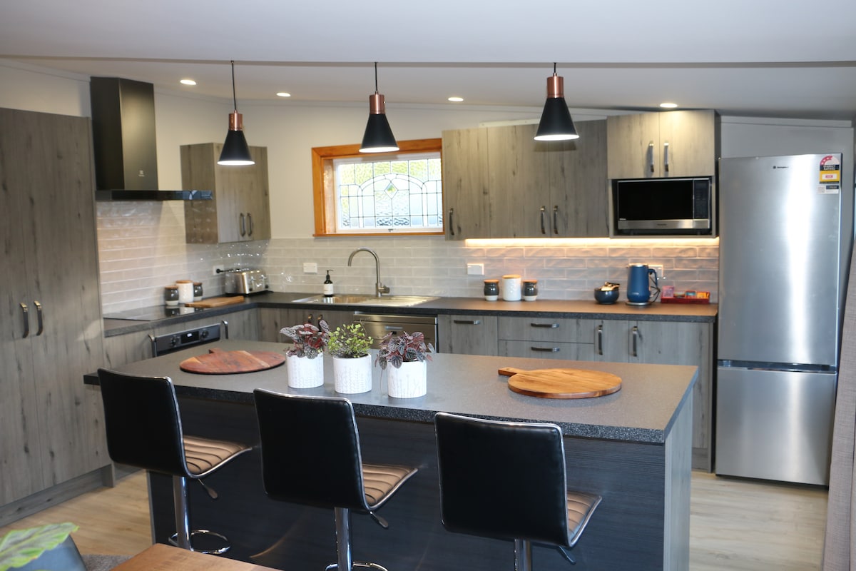 The modern kitchen features sleek cabinetry and warm wood accents. A spacious island with seating is complemented by contemporary pendant lighting. Stainless steel appliances, including a microwave and refrigerator, are visible, along with decorative potted plants enhancing the countertop.