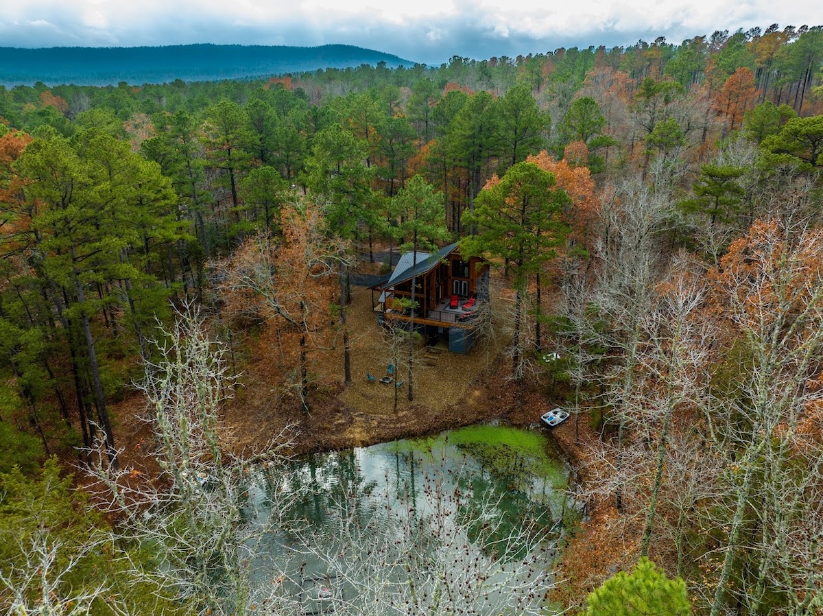 An aerial view showcases a secluded cabin nestled among verdant trees and autumn foliage. A small pond reflects the surrounding natural scenery. The cabin features a modern design with large windows, offering a serene retreat in a wooded area.