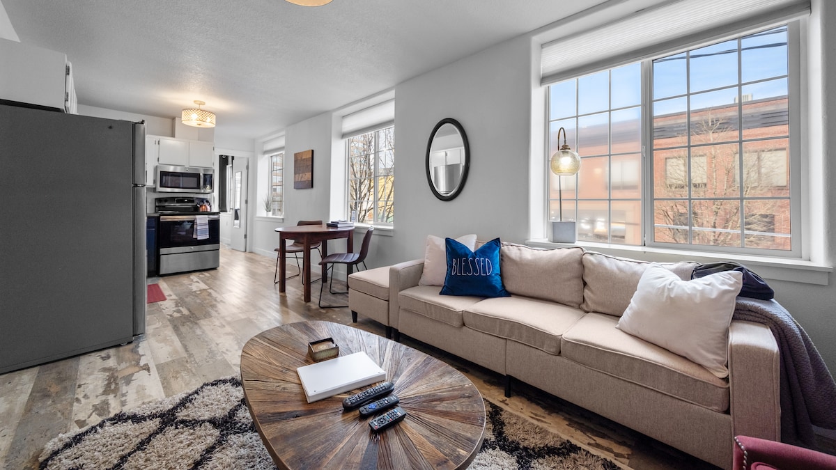 The living area showcases a comfortable sofa adorned with plush pillows, a round wooden coffee table, and a large rug on the floor. Bright light floods the space through two expansive windows, complementing the modern kitchen visible in the background.