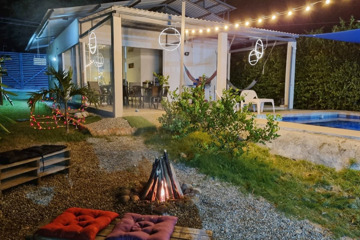 An outdoor area features a fire pit surrounded by colorful cushions on gravel, providing a cozy setting. String lights hang above, illuminating the space. A swimming pool is visible nearby, along with a modern structure that includes outdoor seating and hammocks for relaxation.