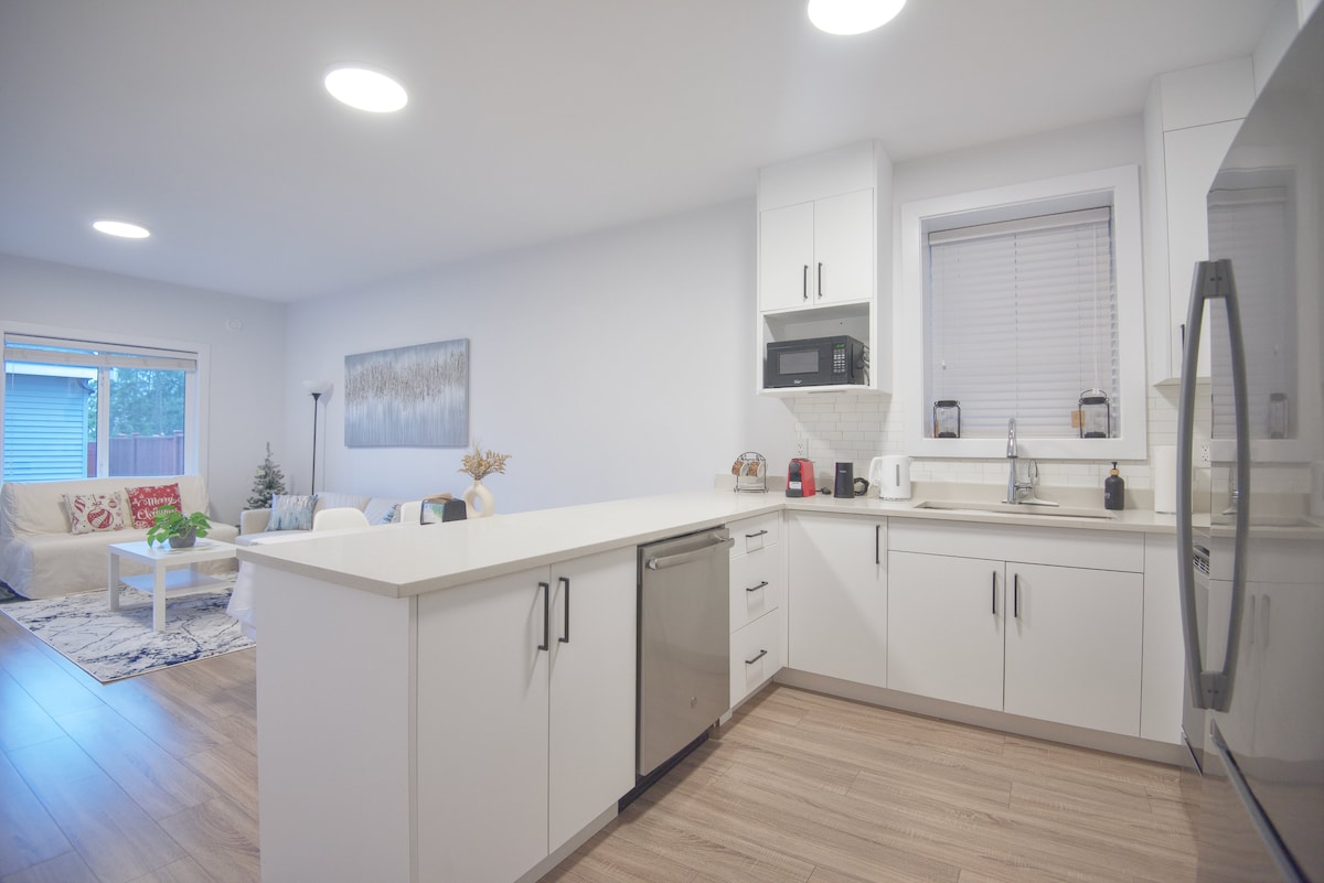 A modern kitchen is presented with sleek white cabinets and a spacious countertop. Stainless steel appliances, including a refrigerator and dishwasher, are visible. Natural light enters through a window, illuminating the open layout that connects to a cozy living area.