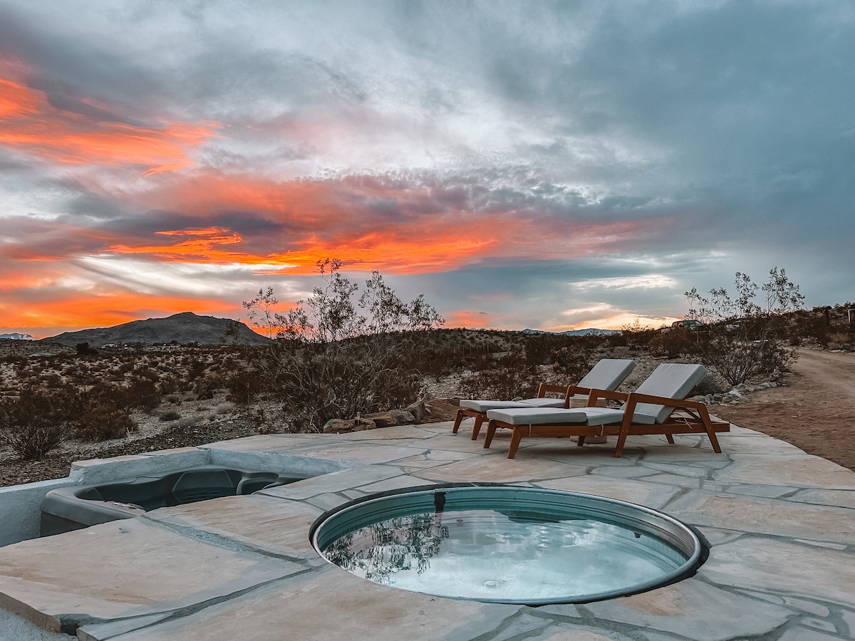A circular soaking tub is set on a stone deck, surrounded by low desert vegetation. Two lounge chairs face the horizon, where a vibrant sunset paints the sky in shades of orange and gray, creating a serene outdoor atmosphere.