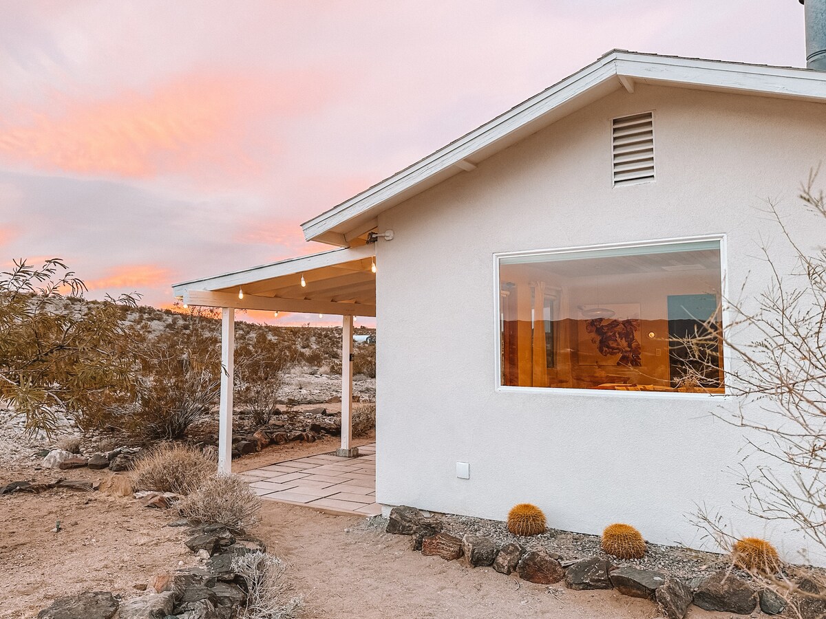 The exterior of the cabin features a light-colored facade with a covered porch, illuminated by string lights. A large picture frame window offers views of the desert landscape, while soft sunset hues fill the sky above.