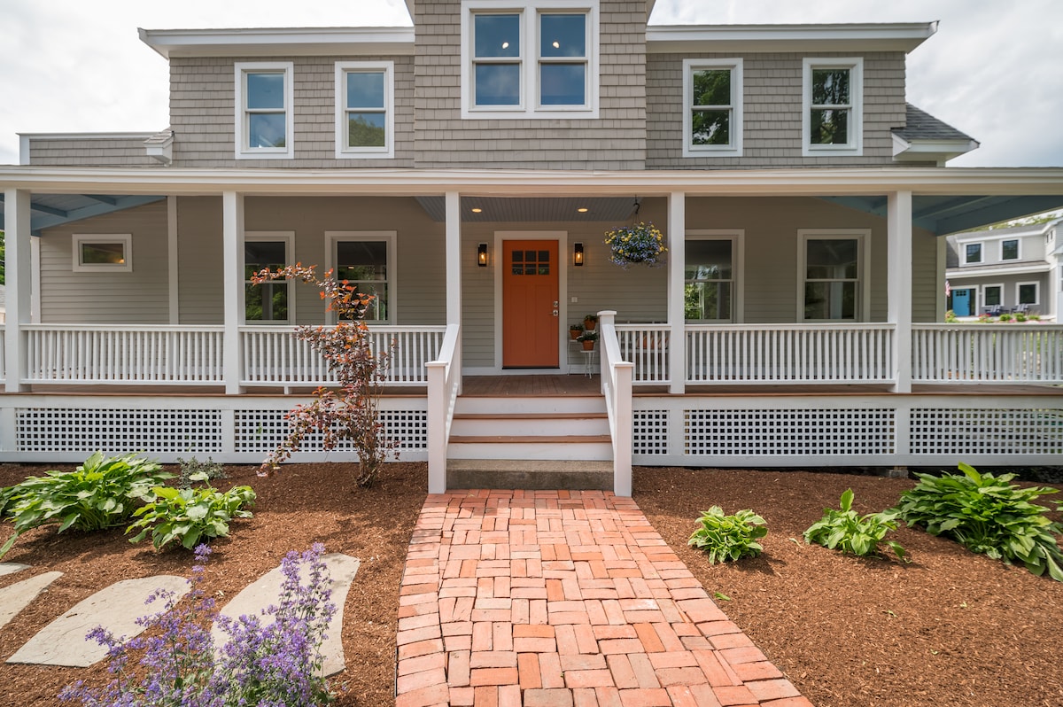 A charming exterior view of a renovated home features a welcoming orange door at the entrance. A spacious covered porch is lined with white railings, complemented by lush landscaping, including purple flowers and greenery. A brick pathway leads to the front steps.
