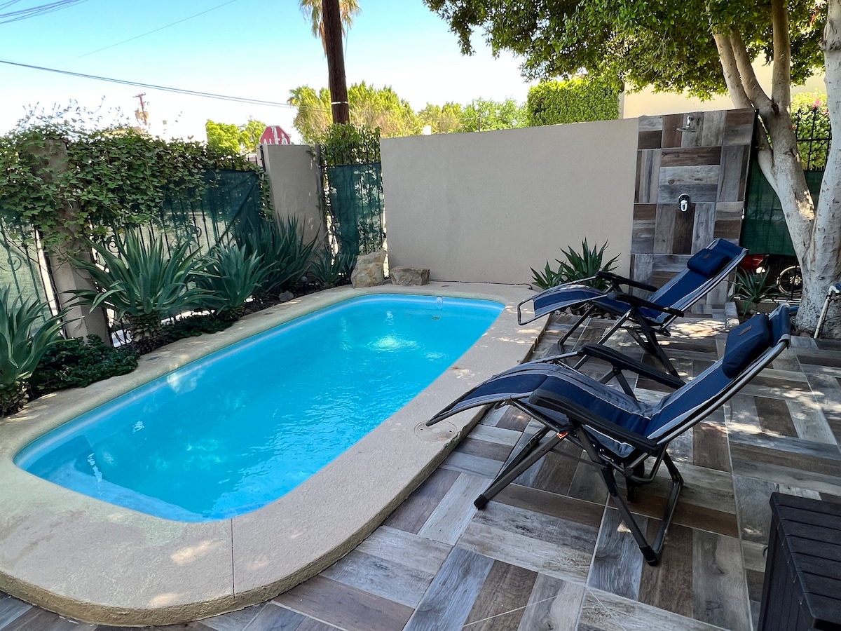 A private outdoor area features a small swimming pool surrounded by lush greenery. Two blue lounge chairs are positioned for relaxation next to the pool. The space has a paved surface with a neutral color scheme, complemented by natural elements.