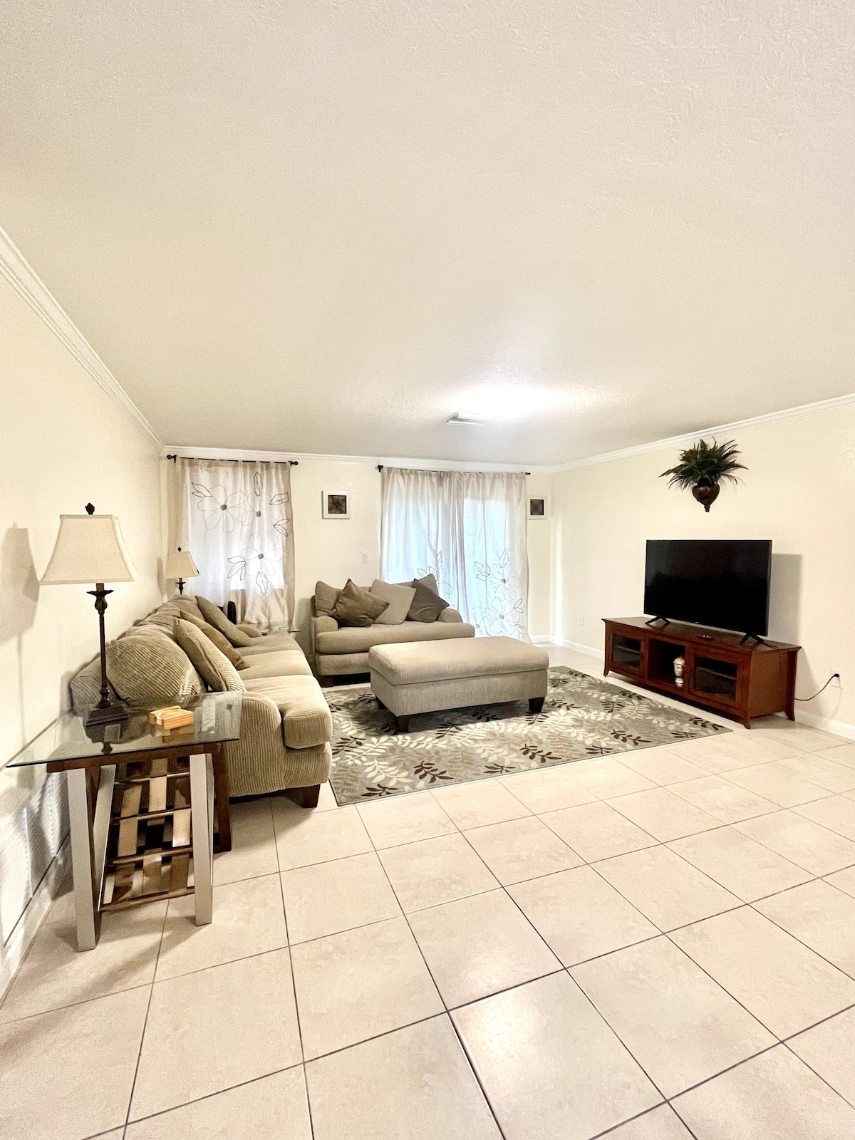 The living room features a comfortable sectional sofa with soft cushions, positioned around a light-colored area rug. Natural light enters through sheer curtains, with a flat-screen TV mounted on the wall opposite a wooden console table. A decorative plant adds greenery to the space.