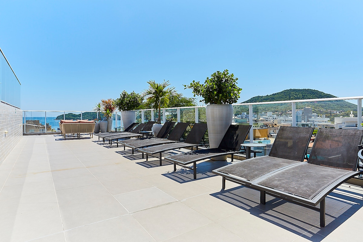 A spacious rooftop area is seen, featuring several lounge chairs arranged around a clean, tiled surface. Potted plants are placed strategically, and the backdrop includes a view of hills and a clear blue sky, creating an inviting environment for relaxation.