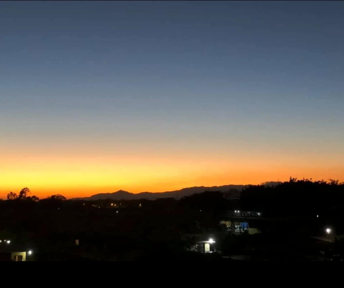 An expansive sky transitions from deep blue to shades of orange and yellow as the sun sets on the horizon. Silhouettes of distant mountains can be seen against the colorful backdrop, with subtle lights from nearby structures dotting the foreground.