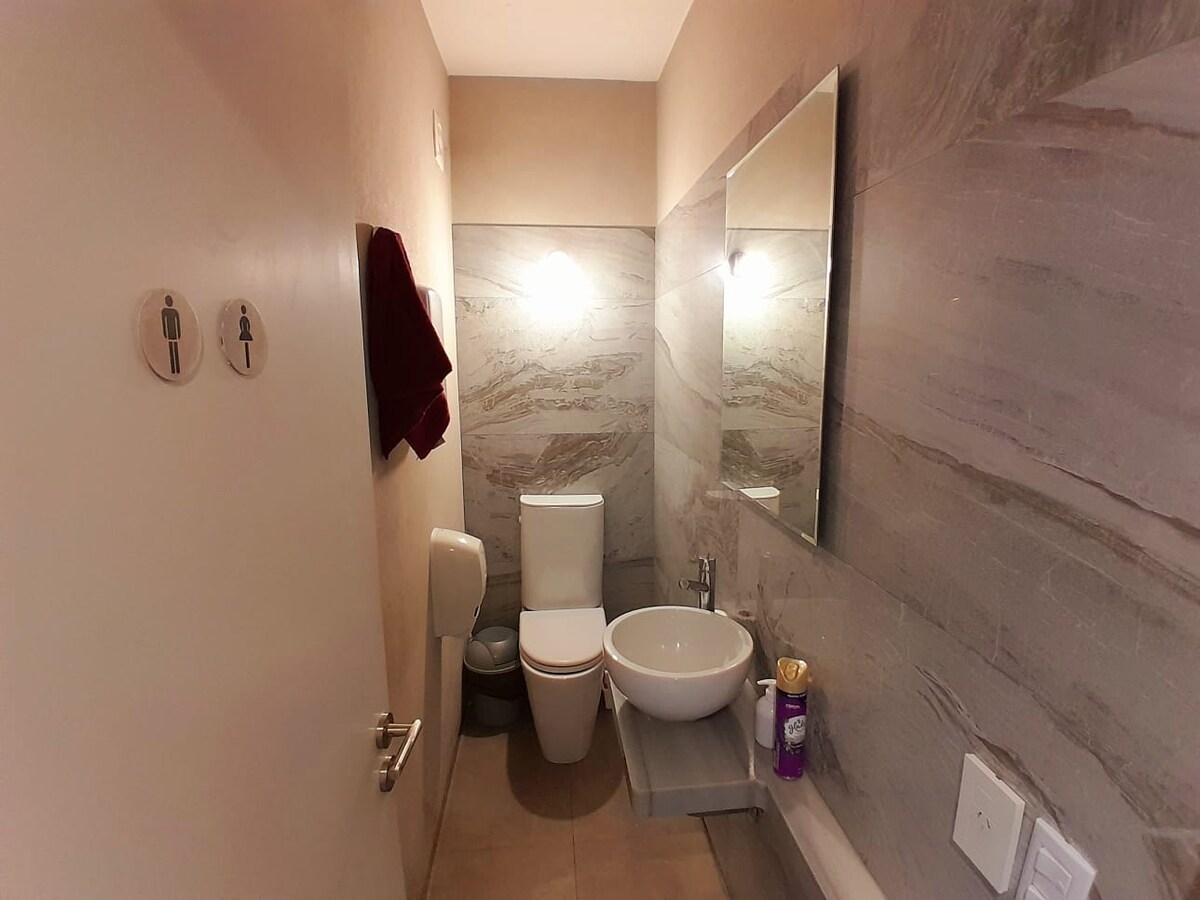 A compact restroom features tasteful grey marble walls. A white toilet and a modern sink are positioned against the wall. A circular mirror reflects soft lighting from above. An accessible grab bar complements the space, while a door leads to a gender-neutral bathroom.