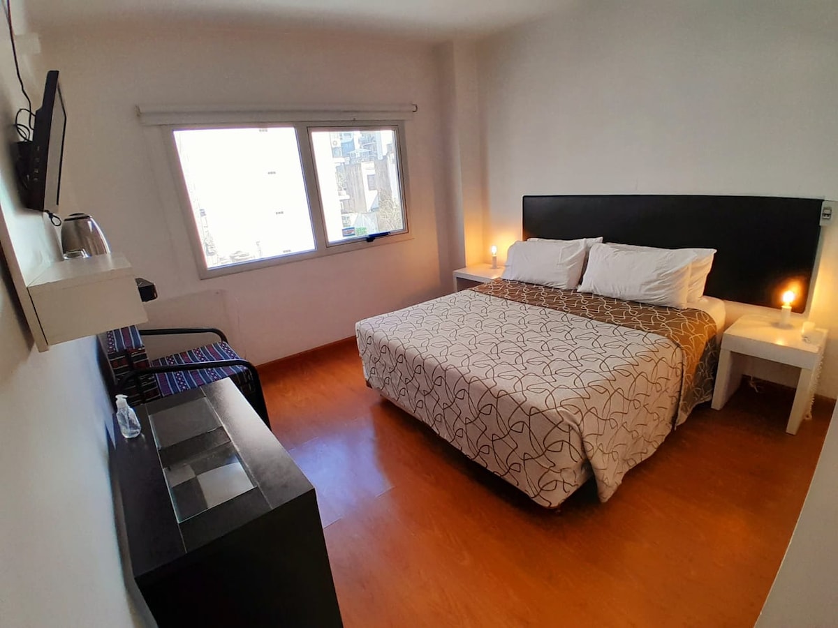 A bright bedroom features a spacious bed with light bedding and a patterned throw. Two nightstands with soft lighting are positioned beside the bed. A flat-screen television is mounted on the wall, and a small table complements a colorful chair in the corner. Natural light fills the room through large windows.