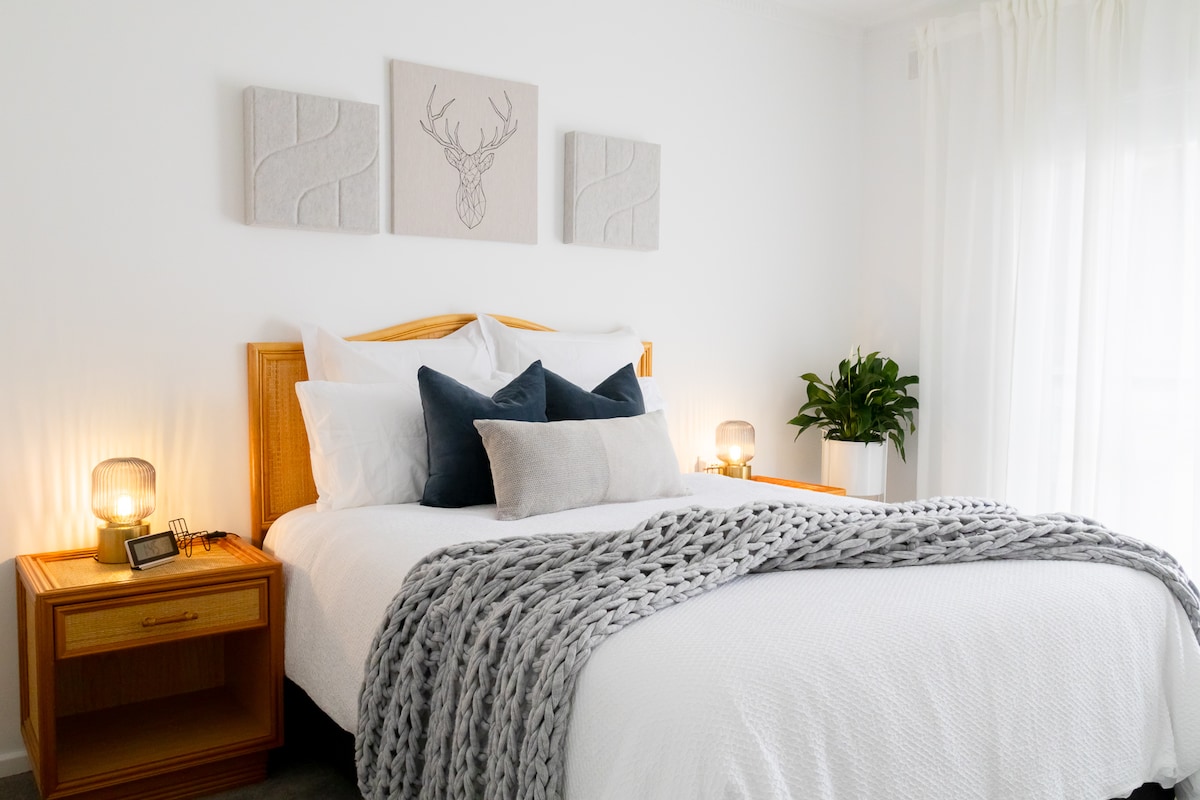 A well-appointed bedroom features a spacious bed adorned with soft white linens and a textured grey throw. Nightstands on either side hold stylish lamps. Above the bed, three minimalist art pieces complement the calming decor. A potted plant adds a touch of greenery to the space.
