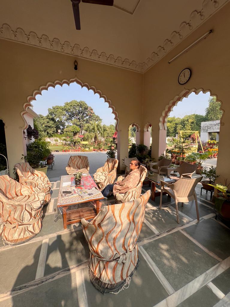 An inviting outdoor seating area features several patterned chairs arranged around a low table. Surrounding plants and vibrant flowers enhance the space. Arched doorways frame the lush garden view, while a clock adorns the wall, adding a functional touch to the serene atmosphere.