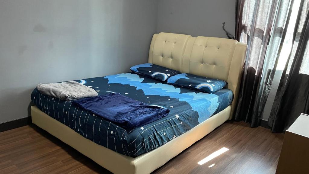A spacious bed with a tufted headboard is centered in the room, dressed in a blue and black geometric patterned coverlet. Soft pillows match the bedding. Natural light enters through a window adorned with sheer and dark curtains, illuminating the warm wood flooring.