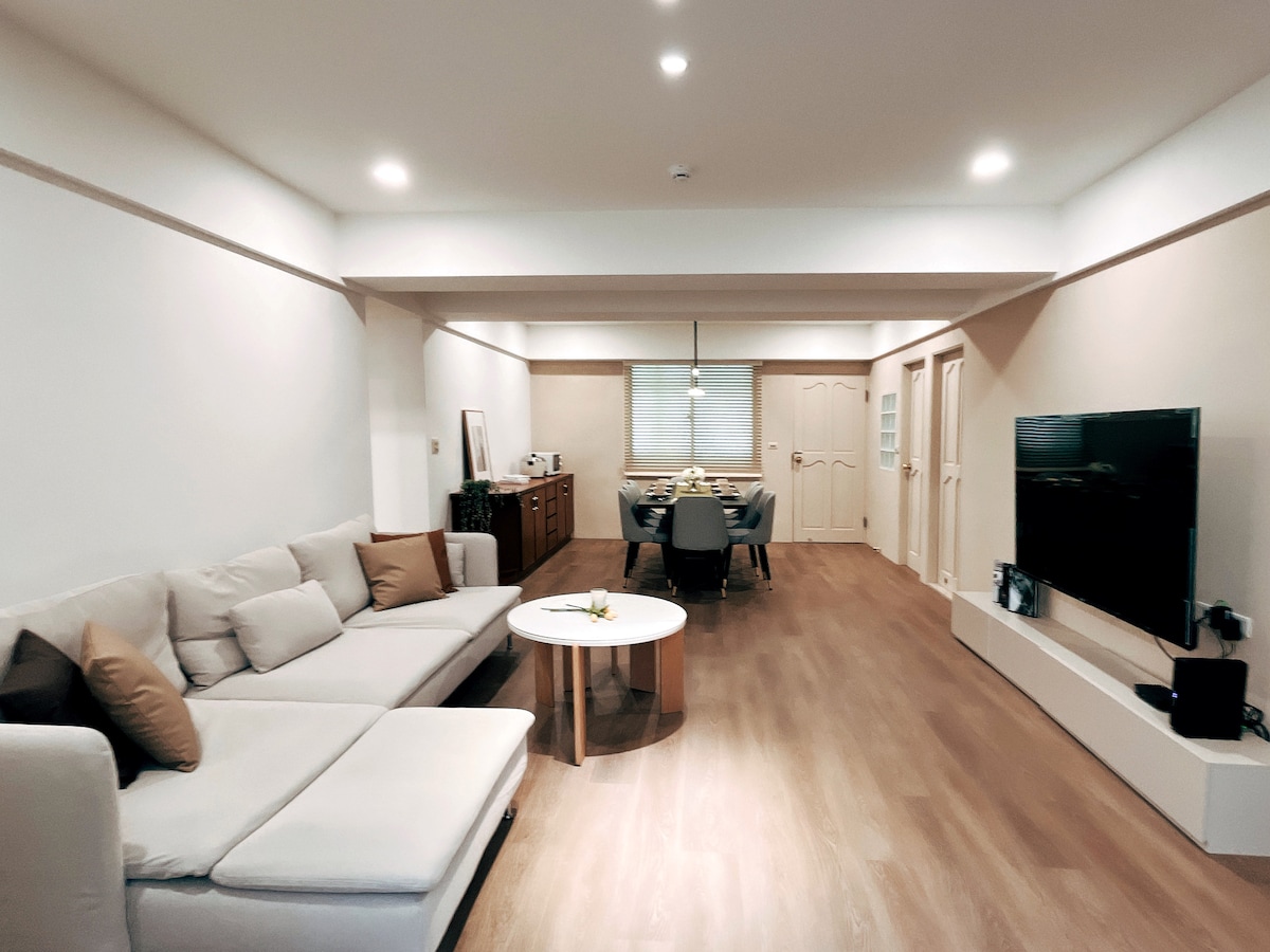 A spacious living area features a sectional sofa with plush cushions, complemented by a coffee table in the center. A large television is mounted on the wall. Light-colored walls and wooden flooring enhance the bright ambiance, with dining space visible in the background.